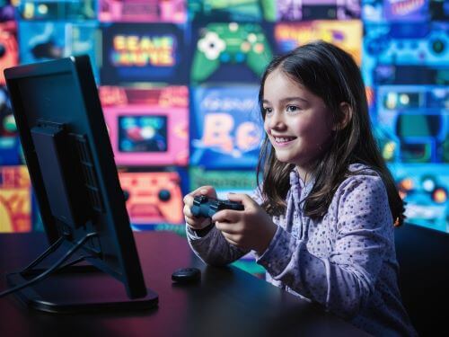 gaming begriffe Madchen sitzt an einem Tisch mit Bildschirm und halt einen Game Controller in der Hand skrivanek