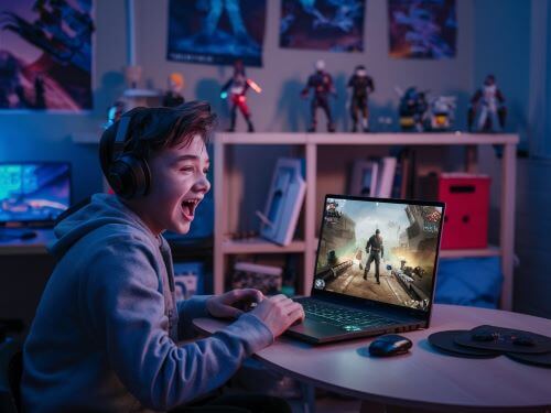 gaming begriffe ein Teenager der an einem Tisch mit einem Laptop sitzt, auf dem ein spannendes Online-Spiel lauft skrivanek